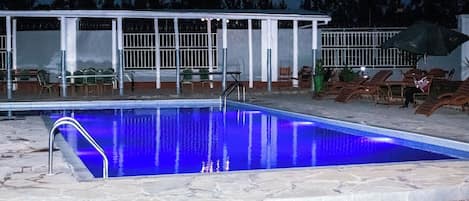 Una piscina de color azul, rodeada de sillones de descanso y una zona de estar cubierta.