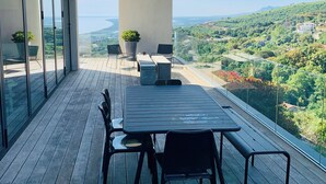 A modern outdoor terrace with a wooden deck, a table with chairs, and a view of a green landscape. - Architect-designed house with sea and mountain views (Bastia)