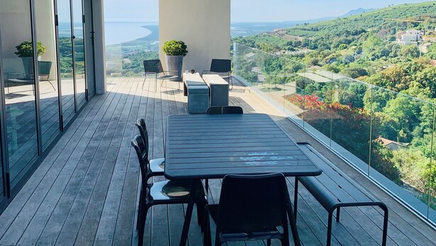 A modern outdoor terrace with a wooden deck, a table with chairs, and a view of a green landscape. - Architect-designed house with sea and mountain views (Bastia)