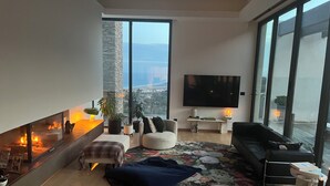 A modern living room with a fireplace, comfortable seating, a large TV, and a view of the city. - Architect-designed house with sea and mountain views (Bastia)