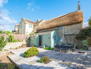 A quaint house with a thatched roof, a stone wall, and a patio with outdoor furniture.