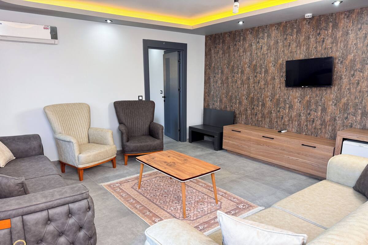 A modern living room with a grey sofa, beige armchair, and a wooden coffee table. A flat-screen TV is mounted on the wall, and a wooden cabinet is against the wall.