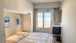 A hotel room with a bed, a small bedside table, a mirror, a window with curtains, and a view of the beach. - Apartment Albertstrand With Stunning Seafront View (Knokke-Heist)
