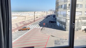 A view from a window overlooking a beach, a road with parked cars, and a building with balconies. - Apartment Albertstrand With Stunning Seafront View (Knokke-Heist)