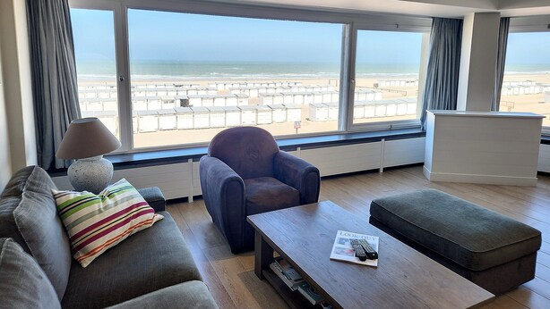 A modern living room with a large window offering a beach view, a sofa with a striped pillow, a dark armchair, and a wooden coffee table with a remote control and magazine. - Apartment Albertstrand With Stunning Seafront View (Knokke-Heist)
