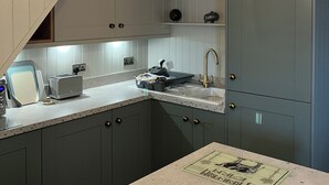 Una cocina con encimera blanca, un lavamanos y un tostador. Hay armarios de tono verdoso y un techo con vigas de madera a la vista.