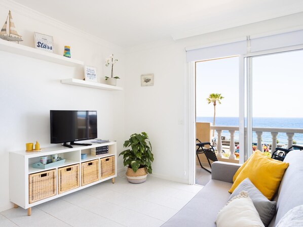 Un salón moderno con televisor, estanterías blancas y un balcón con vistas al mar.