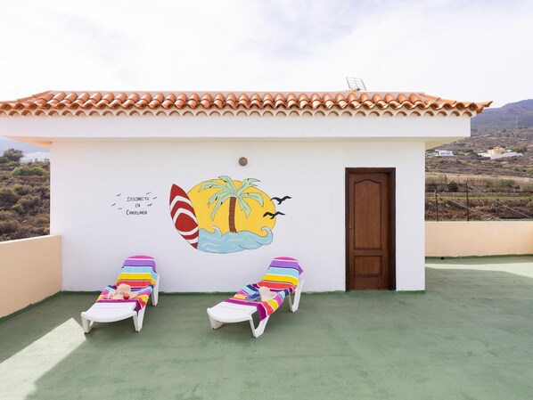 Un petit bâtiment doté d’un toit en tuiles, d’une fresque représentant une scène de plage et de deux chaises longues avec des coussins rayés aux couleurs vives.