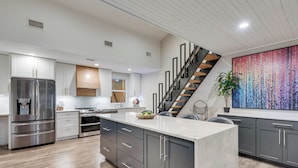 A modern kitchen with a central island, stainless steel appliances, grey cabinets, and a wooden staircase.