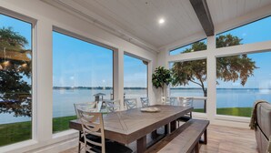 A spacious dining area with a large wooden table and chairs, large windows offering a view of a lake and trees.