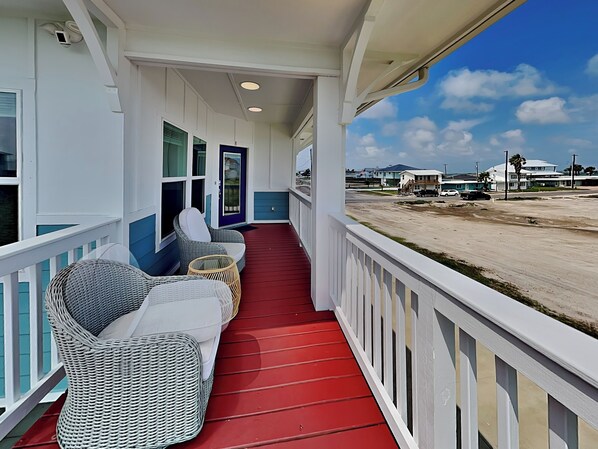 A covered porch with wicker furniture and a red floor, overlooking a beach and residential area. - Inn at Turtle Cove | Bay Views + Pool! (Rockport)