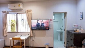 A room with a TV displaying a show, a desk with a chair, a window with curtains, a door leading to a bathroom, and a small trash bin. - Seven Rabbit House (Bangkok)
