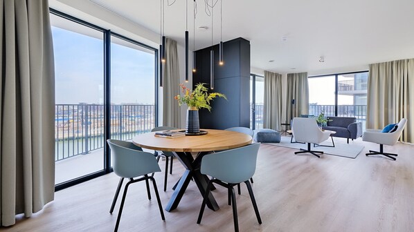 A modern living room with a round wooden table, two blue chairs, a grey sofa, and a white armchair. There is a large window with a view of a cityscape. - Dormio Breskens Apartments & Penthouses (Breskens)