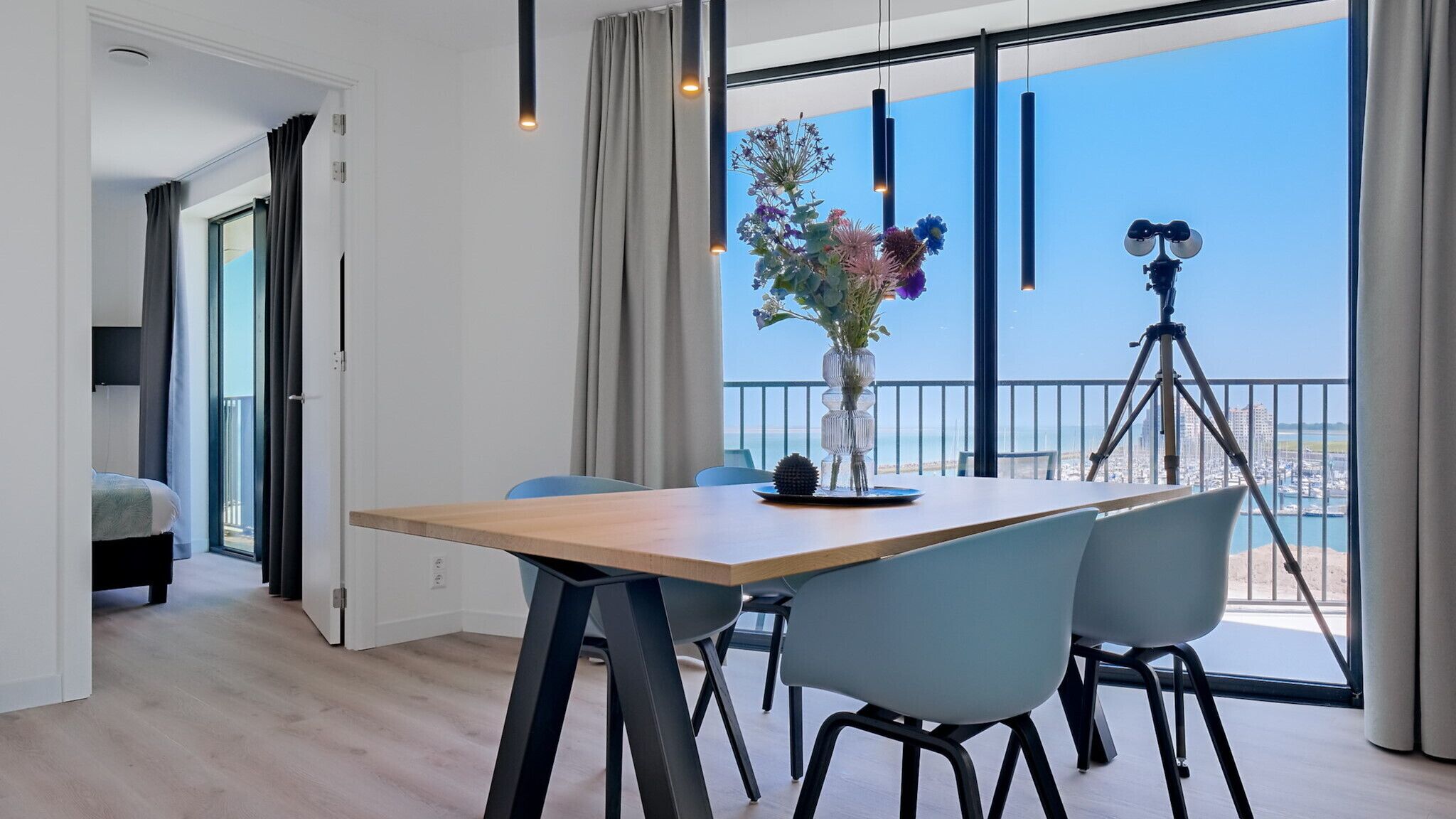 A modern dining area with a wooden table and chairs, a vase with flowers, and a balcony with a view of the city and sea.