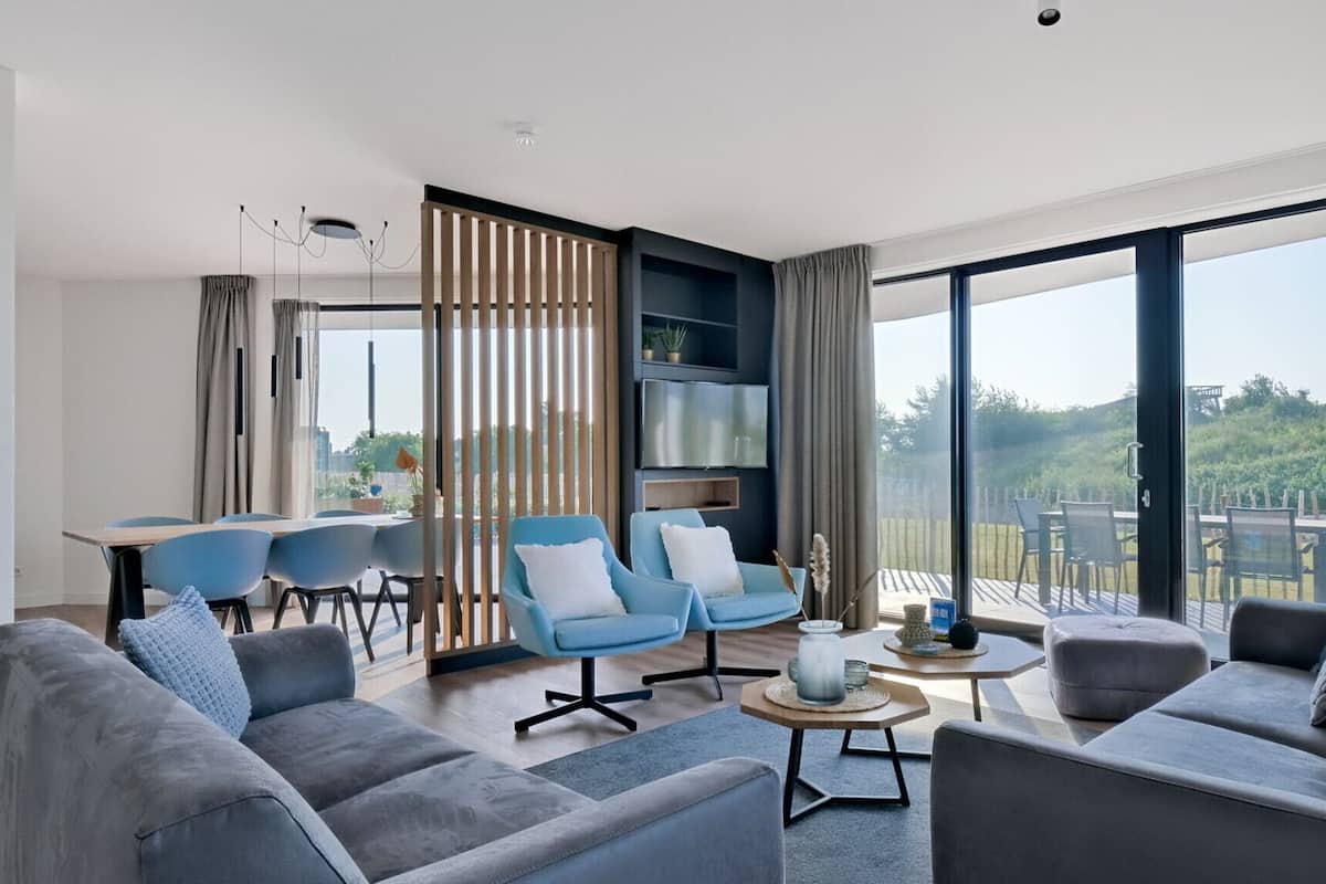 A modern living room with grey sofas, blue armchairs, and a dining area with a table and chairs. Large windows offer a view of greenery outside.