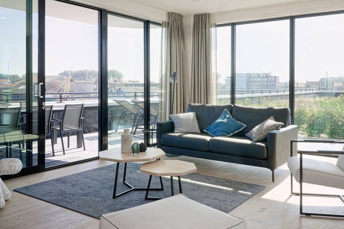 A modern living room with a sofa, coffee table, and chairs, large windows with a city view, and a glass door leading to an outdoor area.