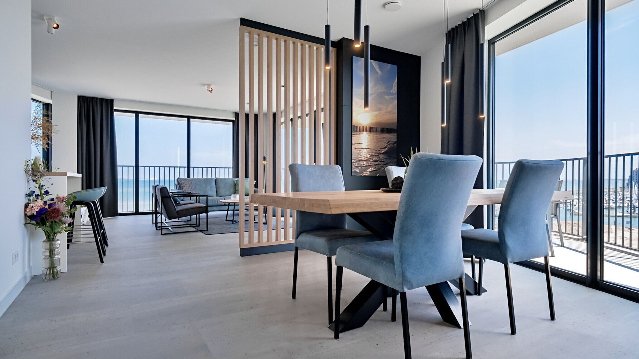 A modern dining area with a wooden table and blue chairs, large windows with a view of the sea, and a decorative plant on the left.