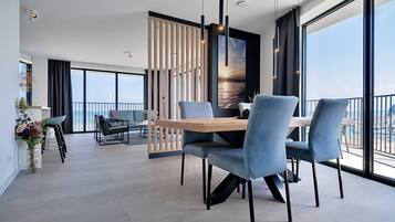 A modern dining area with a wooden table and blue chairs, large windows with a view of the sea, and a decorative plant on the left.