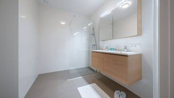 Un baño moderno con cabina de ducha de vidrio, tocador de madera con encimera blanca y un espejo grande sobre el lavabo.