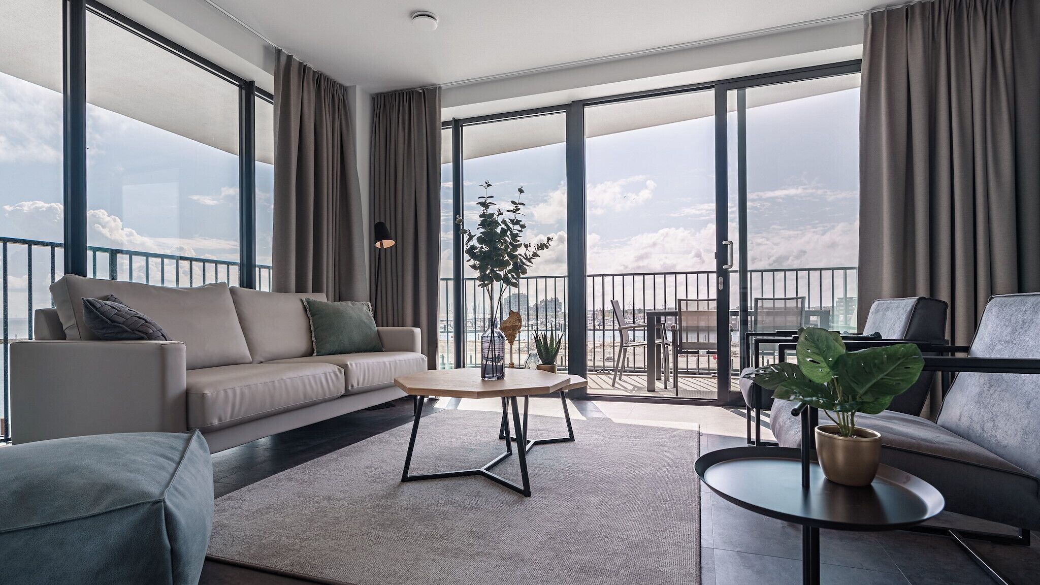 A modern living room with a sofa, coffee table, and chairs, large windows with curtains, and a balcony with a view.