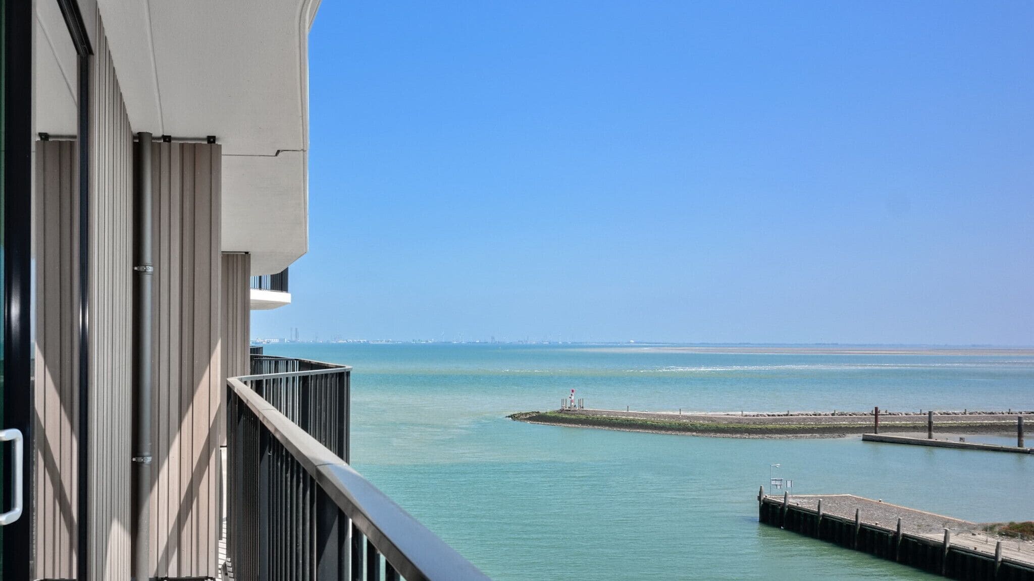 Un balcon offrant une vue sur une mer calme, les contours d’une ville lointaine et une petite île surmontée d’un phare.