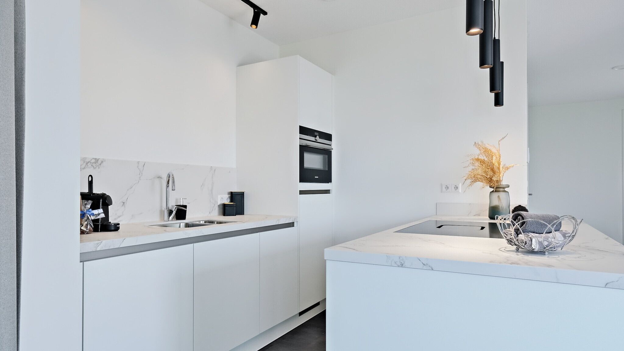 Une cuisine moderne avec des meubles blancs, un plan de travail en marbre, un four encastré et un petit îlot central agrémenté d’un vase contenant des plantes séchées.