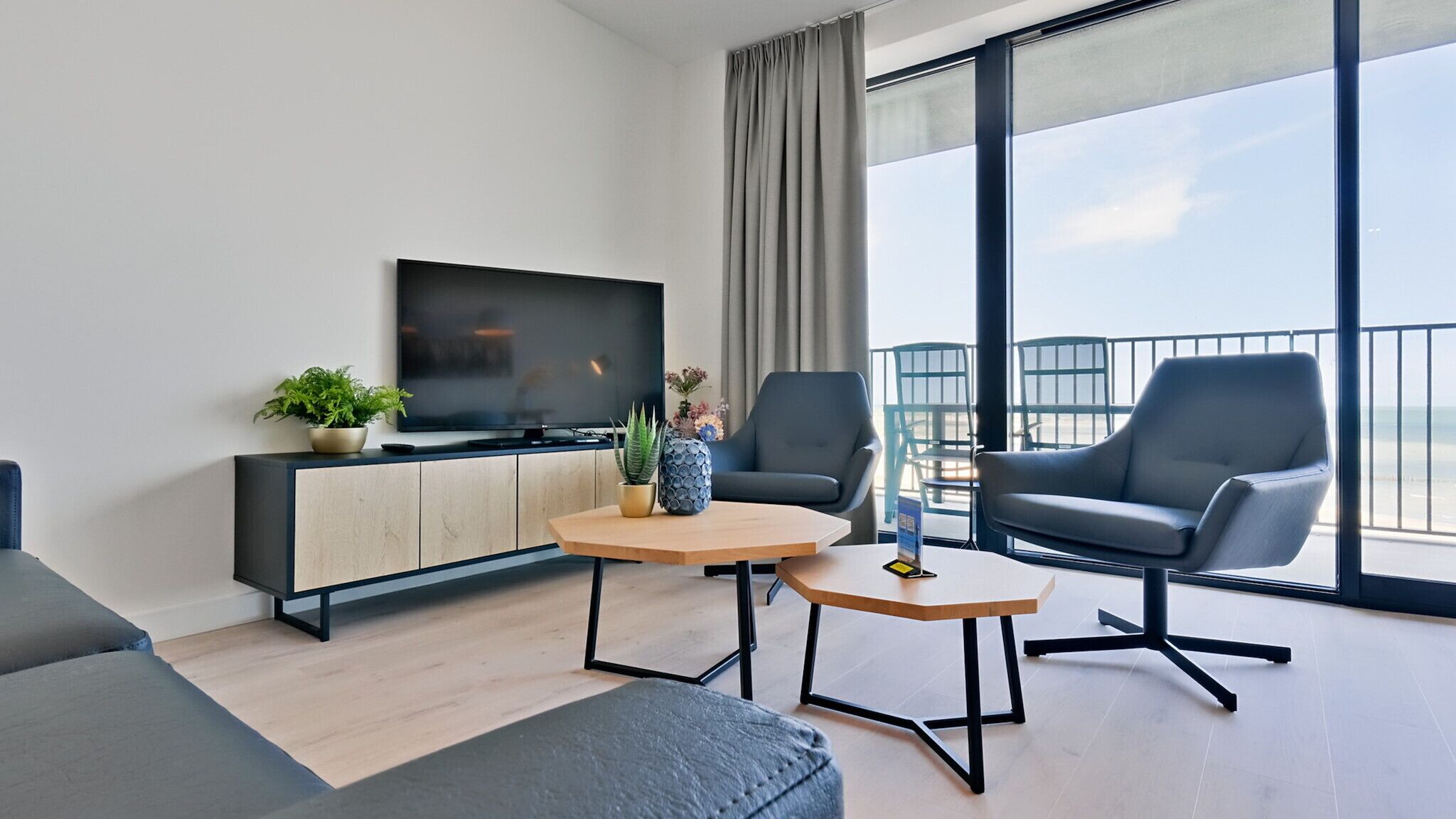 A modern living room with a flat-screen TV, two armchairs, a sofa, a coffee table, and a balcony with a view.