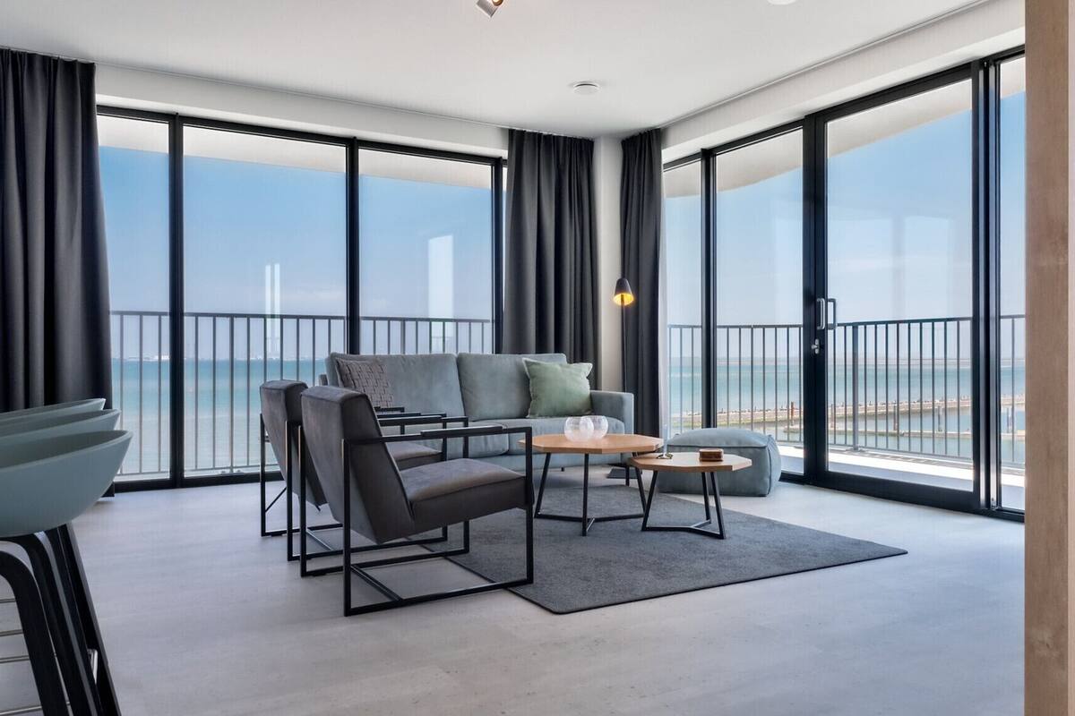 A modern living room with a sofa, two armchairs, a coffee table, and a view of the sea through large windows.