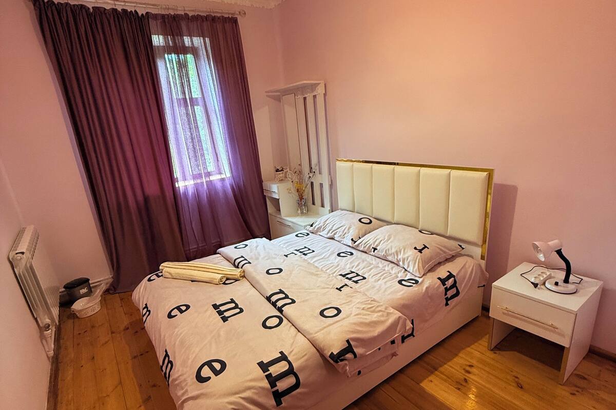 A bedroom with a bed, a nightstand, a lamp, a window with curtains, and a ceiling light.