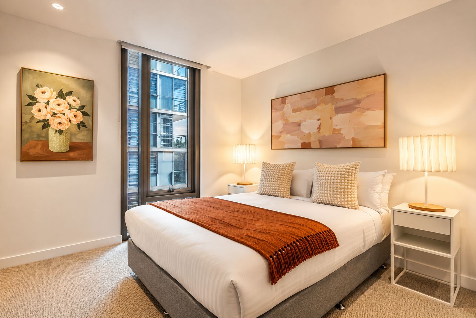 A bedroom with a large bed, two bedside tables with lamps, a window with blinds, and artwork on the walls.