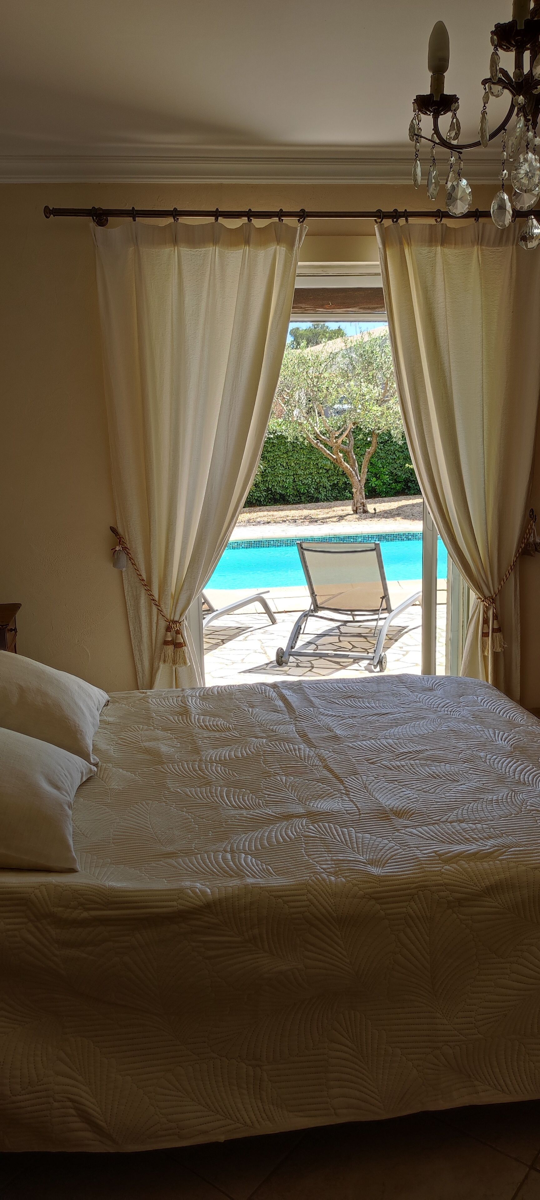 A bedroom with a large bed, a view of a pool and a chair outside, and a chandelier.