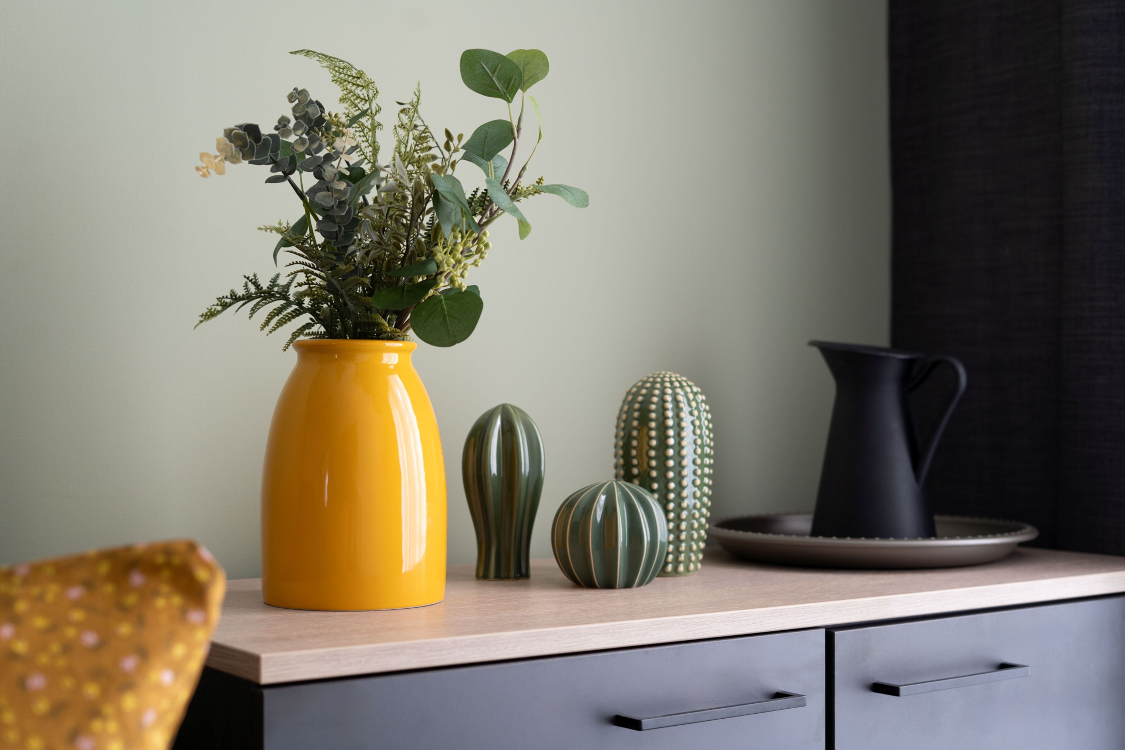 Una encimera de cocina con un florero amarillo que contiene una planta, dos floreros verdes y una jarra negra.