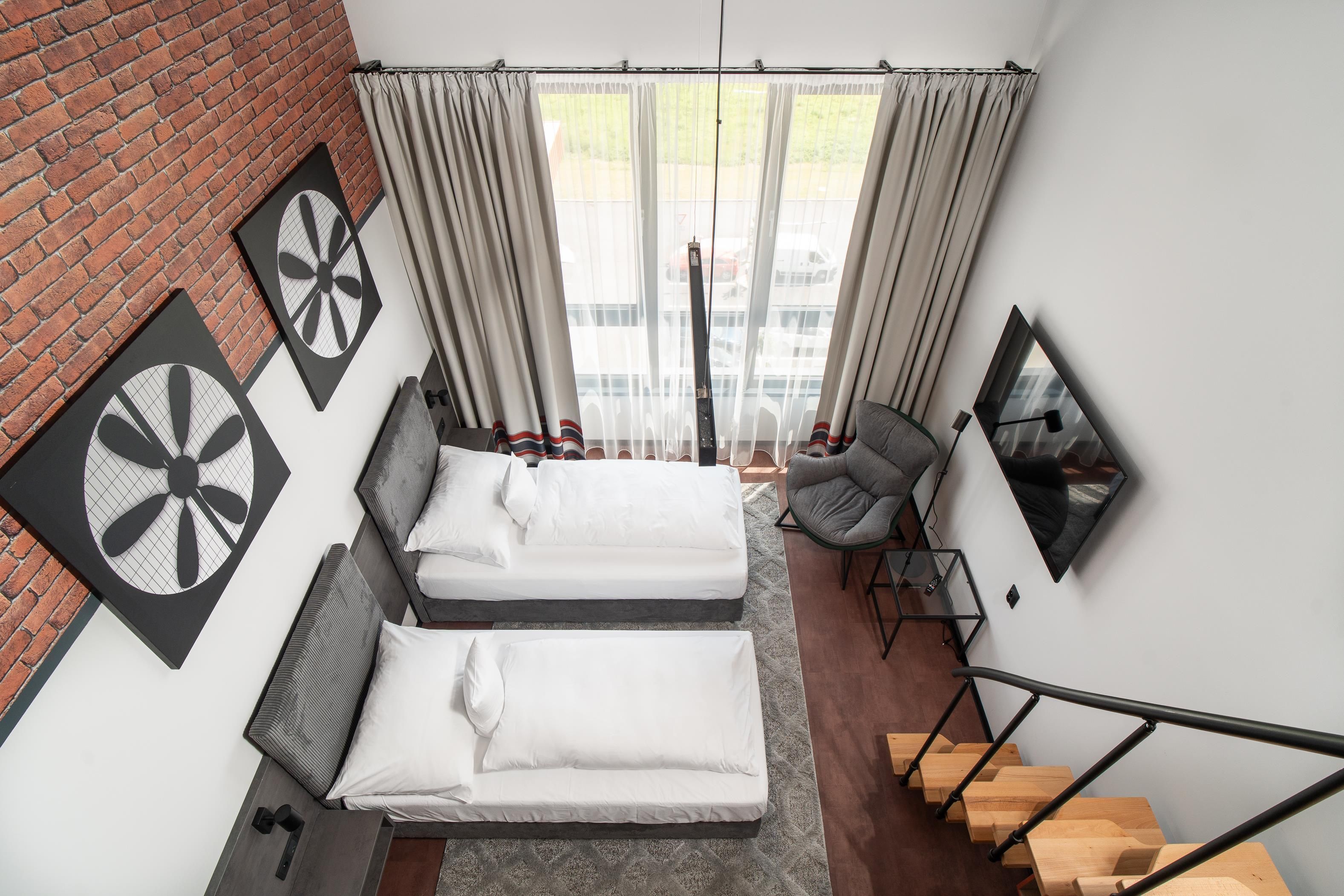 Quarto de hotel moderno com duas camas, uma parede de tijolos, uma janela grande com cortinas, uma cadeira, uma escrivaninha e uma escada.