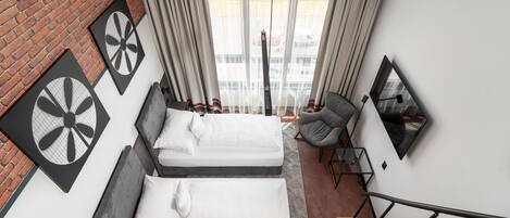 A modern hotel room with two beds, a brick wall, a large window with curtains, a chair, a desk, and a staircase.