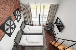 Habitación de hotel moderna con dos camas, una pared de ladrillo, un ventanal con cortinas, una silla, un escritorio y una escalera.