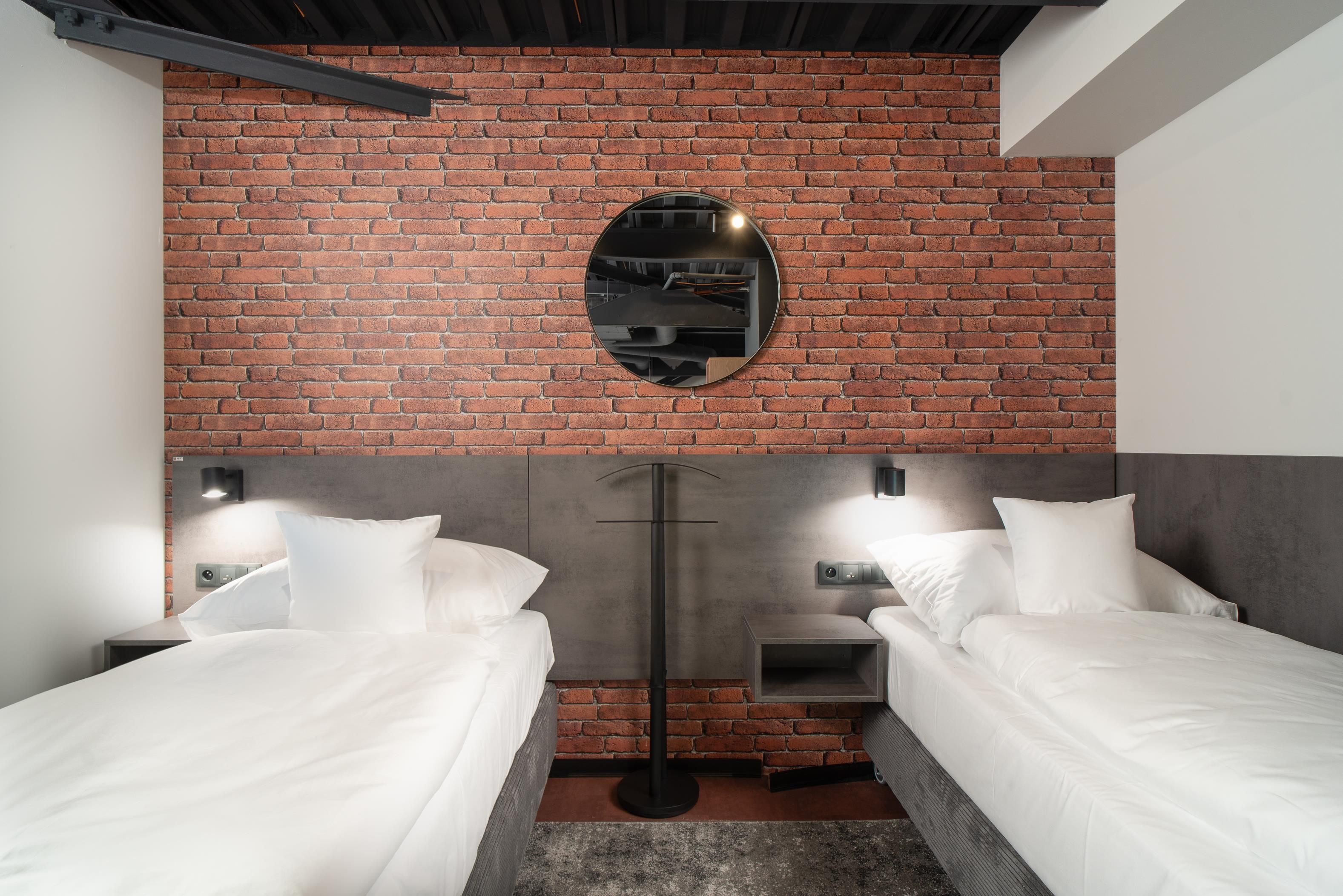 Habitación de hotel con dos camas, una pared de ladrillo, un espejo redondo y un cabecero moderno.