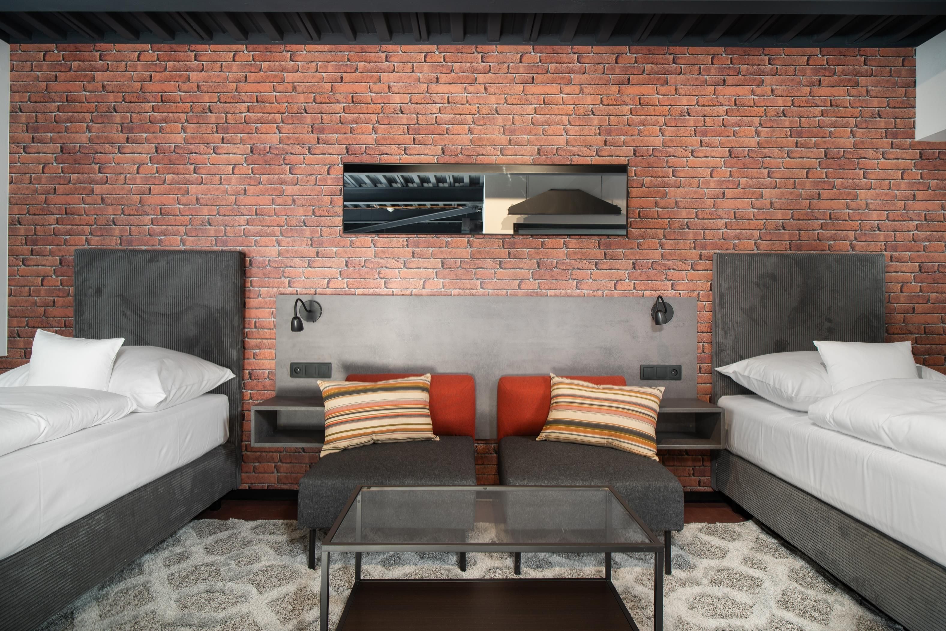 A hotel room with a brick wall, two beds, a sofa, a glass coffee table, and a small TV mounted above the sofa.