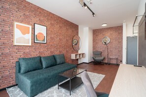 Una sala de estar moderna con pared de ladrillo, un sofá verde, una mesa de centro de cristal y dos relojes en la pared.