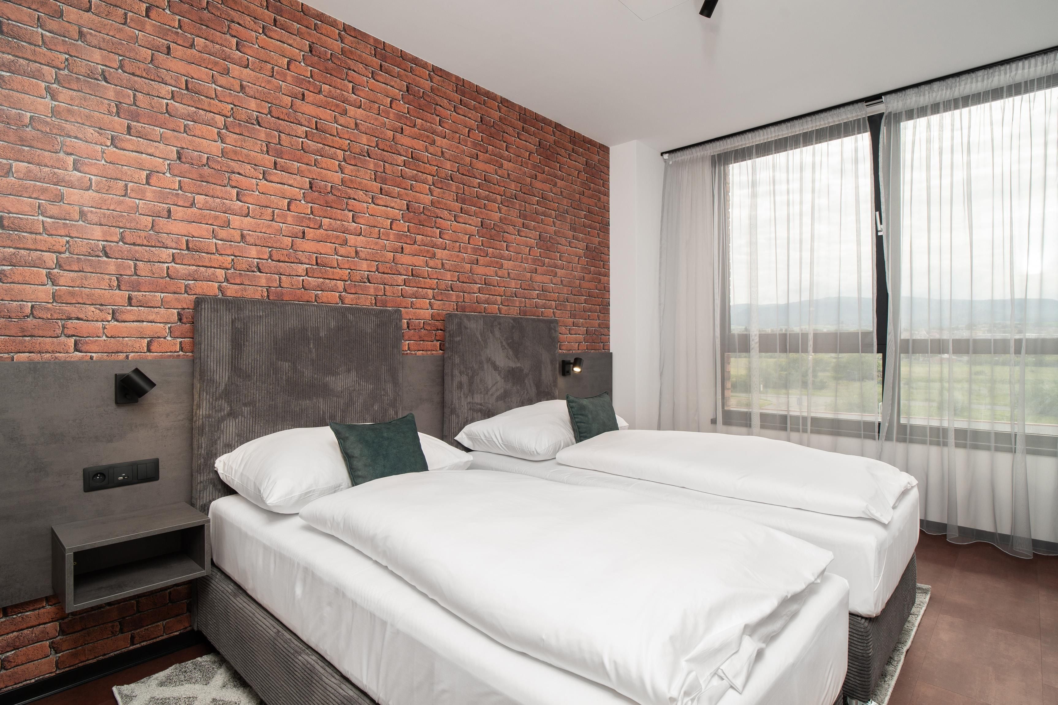 A hotel room with a brick wall, two beds, a nightstand, a wall-mounted light, and a large window with sheer curtains.
