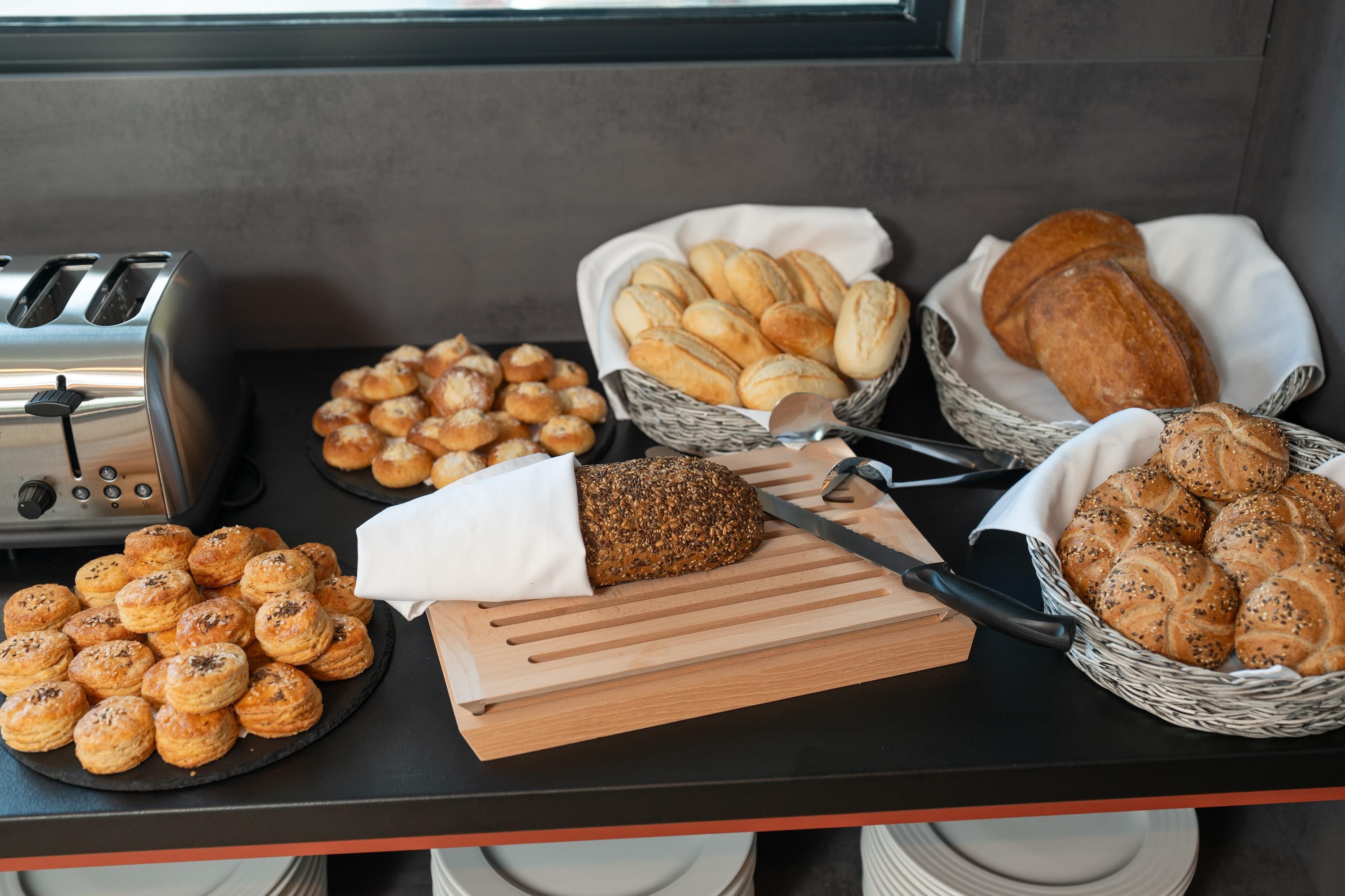 A buffet table with various breads and pastries, a toaster, and a cutting board.
