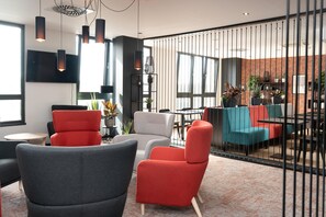 Un espace de détente moderne offrant de nombreuses options d’assises, notamment des fauteuils rouges et gris, ainsi qu’un mur de briques agrémenté de grandes fenêtres.