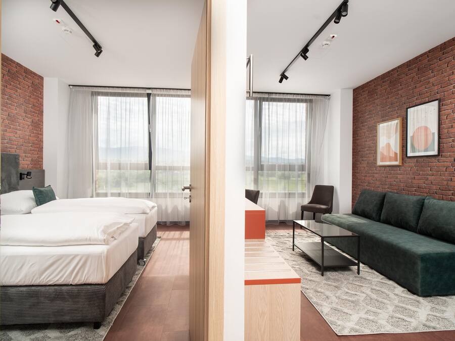 A modern hotel room with a brick wall, two beds, a green sofa, and a large window.