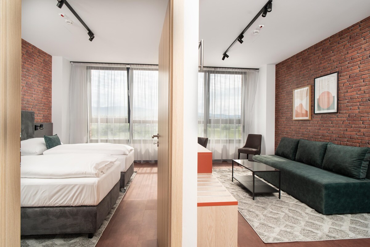 A modern hotel room with a brick wall, two beds, a green sofa, a coffee table, and large windows with sheer curtains.