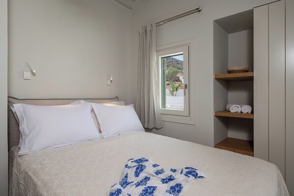 A neatly made bed with a white and blue patterned blanket, a wooden headboard, a window with a view of the outside, and a shelf with towels and a basket. - Castello Amorgos Suites (Amorgos)