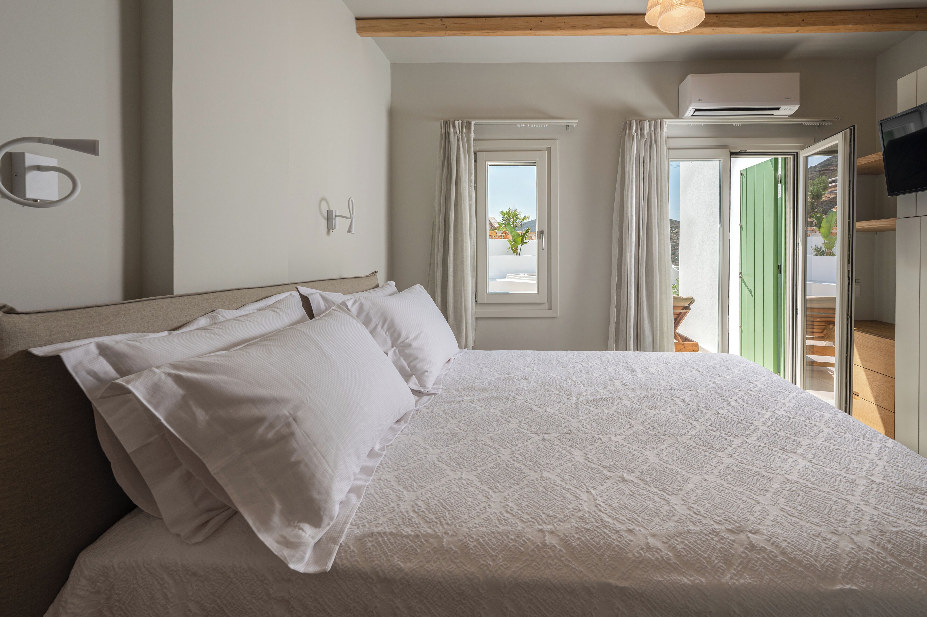A neatly made bed with white linens in a room with a wooden ceiling, a wall-mounted light fixture, and a window with a view of greenery.