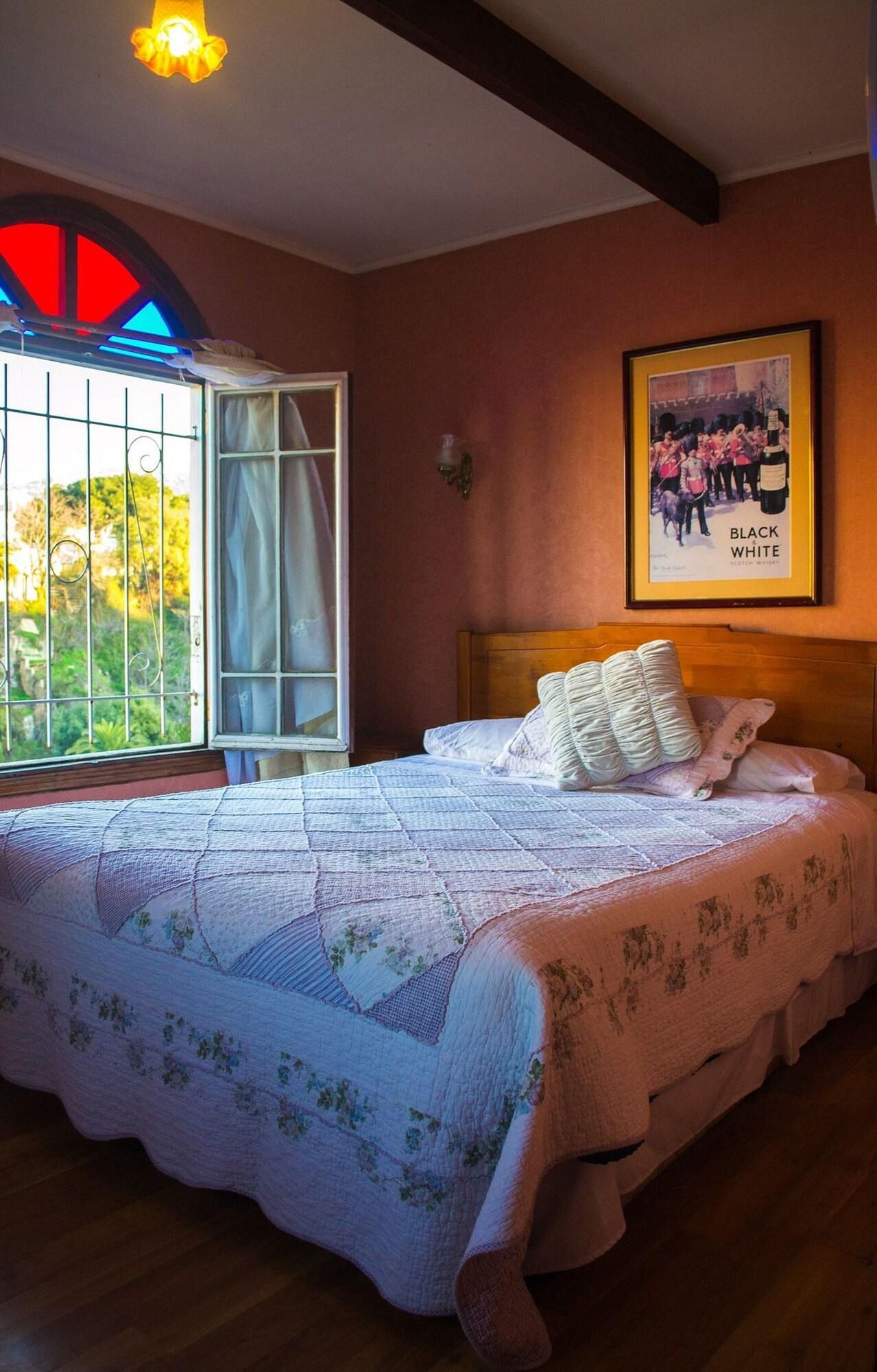 A bedroom with a large bed, a window with a view of greenery, and a framed poster on the wall.
