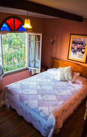 A bedroom with a bed, a window with a view of greenery, a framed picture on the wall, and a hanging light fixture. - Hotel Brighton (Valparaiso)