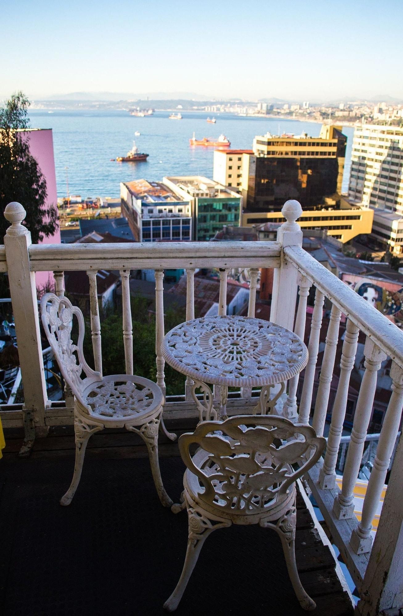 Un balcón con sillas y mesa de metal blanco ornamentado, con vistas a un paisaje urbano y una masa de agua.