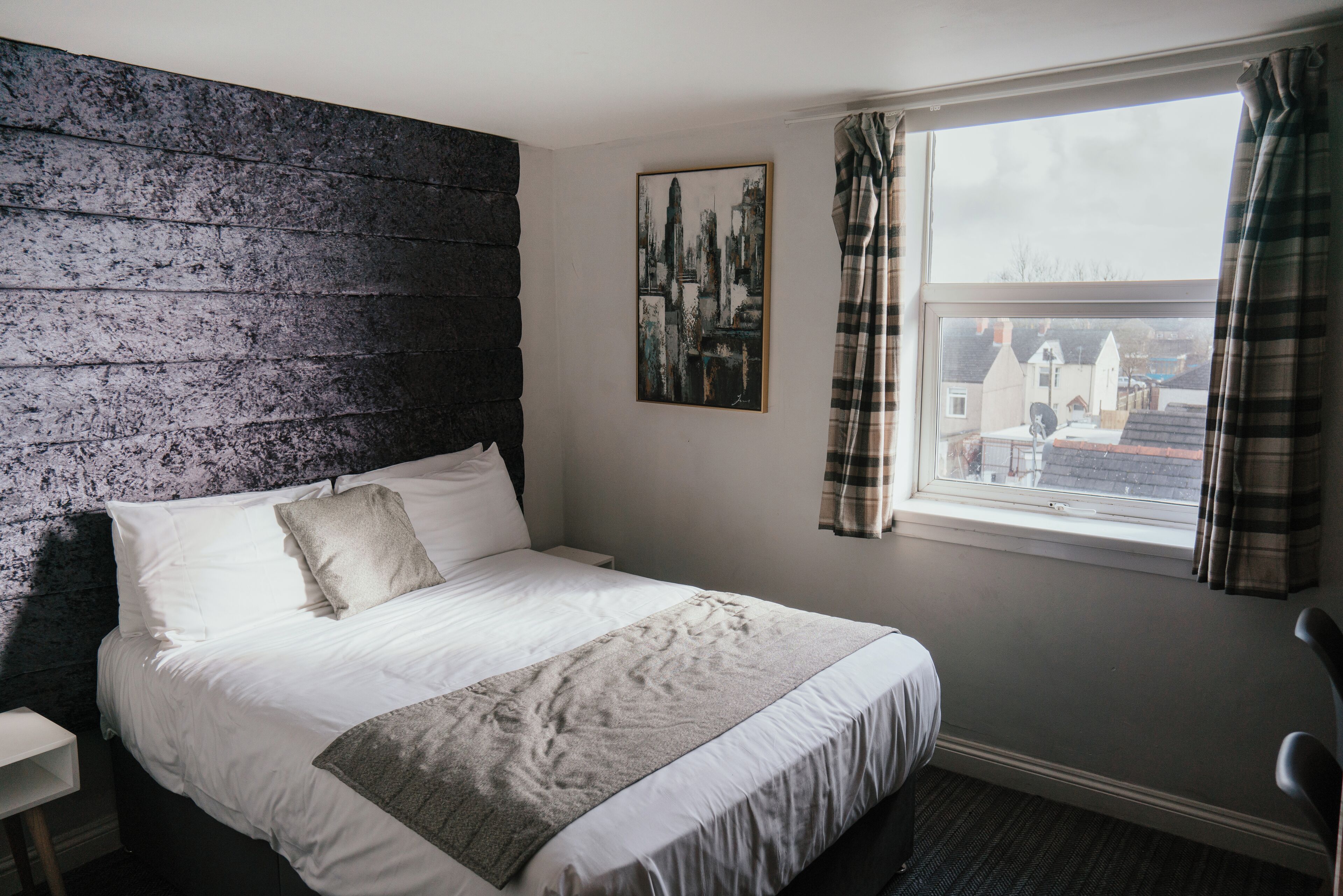 A bedroom with a bed, a headboard, a window with curtains, a wall-mounted picture, and a small bedside table.