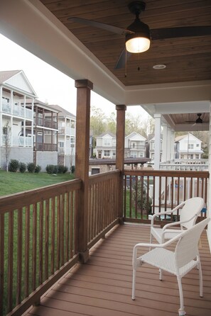 A covered outdoor area with a wooden deck, white wicker chairs, and a view of multi-story residential buildings.